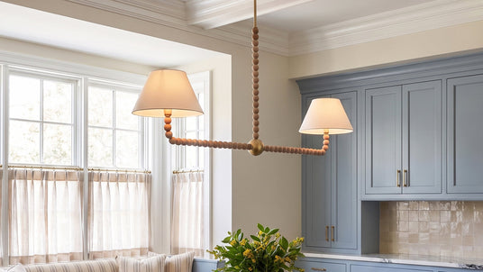 The Casimir Linear Pendant features natural wooden beads and a Brass finished with white fabric shades. Shown in a eat-in kitchen with French blue cabinets and a coffered ceiling.