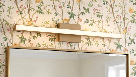 A Garrison LED Vanity Light 30", Satin Brass finish shown above a vanity mirror with colorful floral wallpaper.