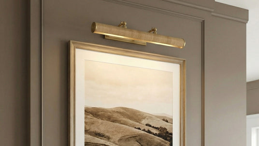 Judy Large Picture Light 24", Aged Brass and Grasscloth shown illuminating a sepia toned photography in a gold frame