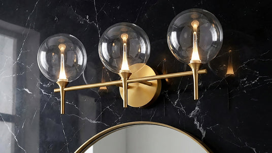 Eaton LED 3 Light Vanity, Brass and Clear Glass finish, on a black marble bathroom wall.