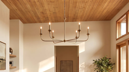 The Harper 6 Light 49" wide Chandelier in Aged Brass finish hung in a modern living room with vaulted natural wood ceiling with wooden window frames, and warm afternoon light streaming in.