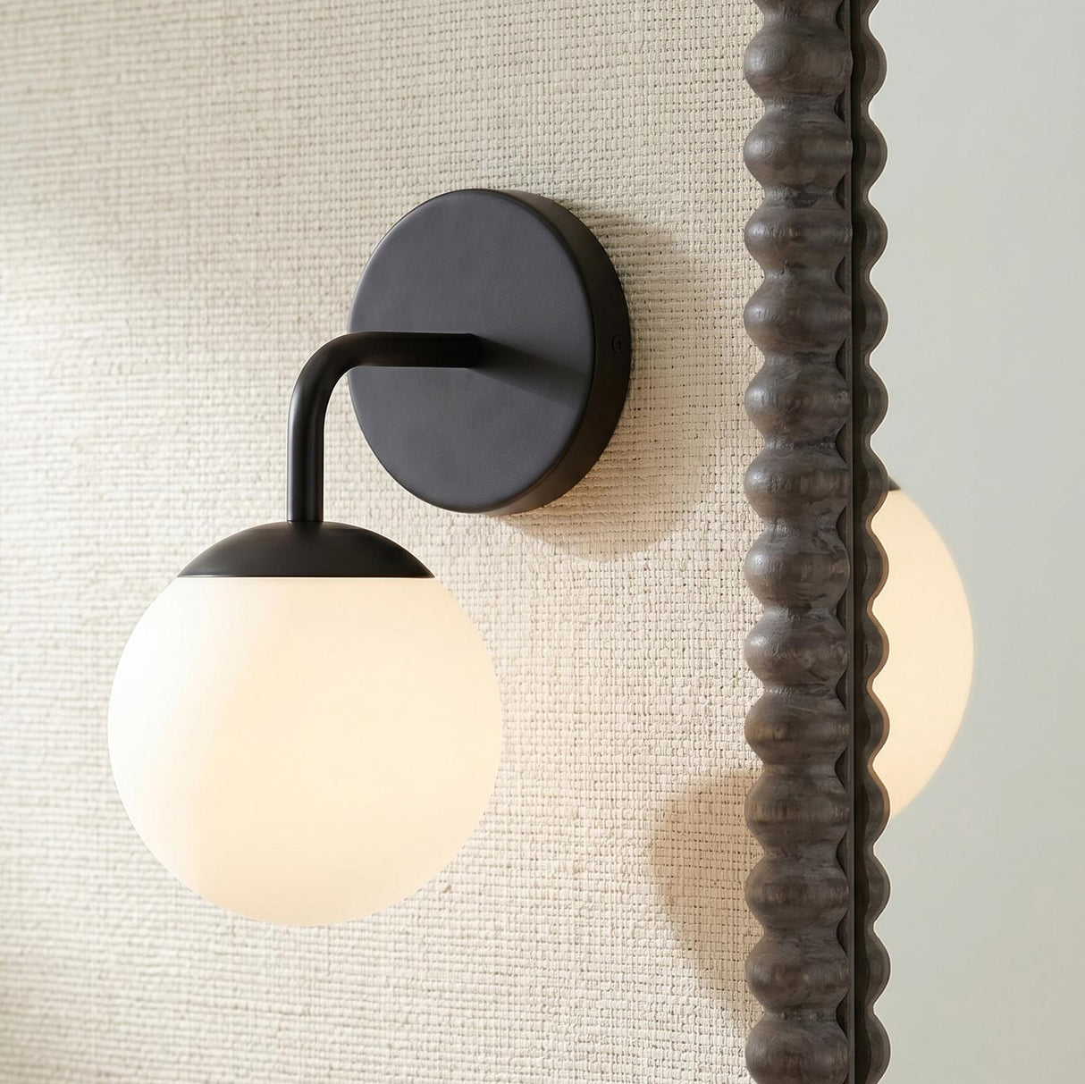 A modern bathroom with the Castell Single Globe Vanity Light, Matte Black featuring a frosted glass globe mounted on a beige textured wall beside a dark bobbin mirror.
