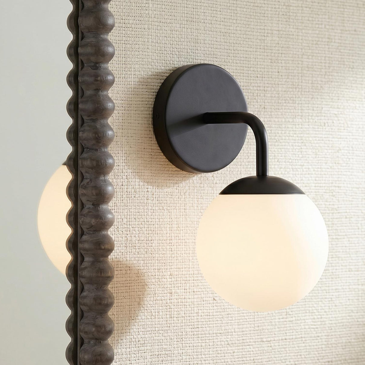 A modern bathroom with the Castell Single Globe Vanity Light, Matte Black featuring a frosted glass globe mounted on a beige textured wall beside a dark bobbin mirror.