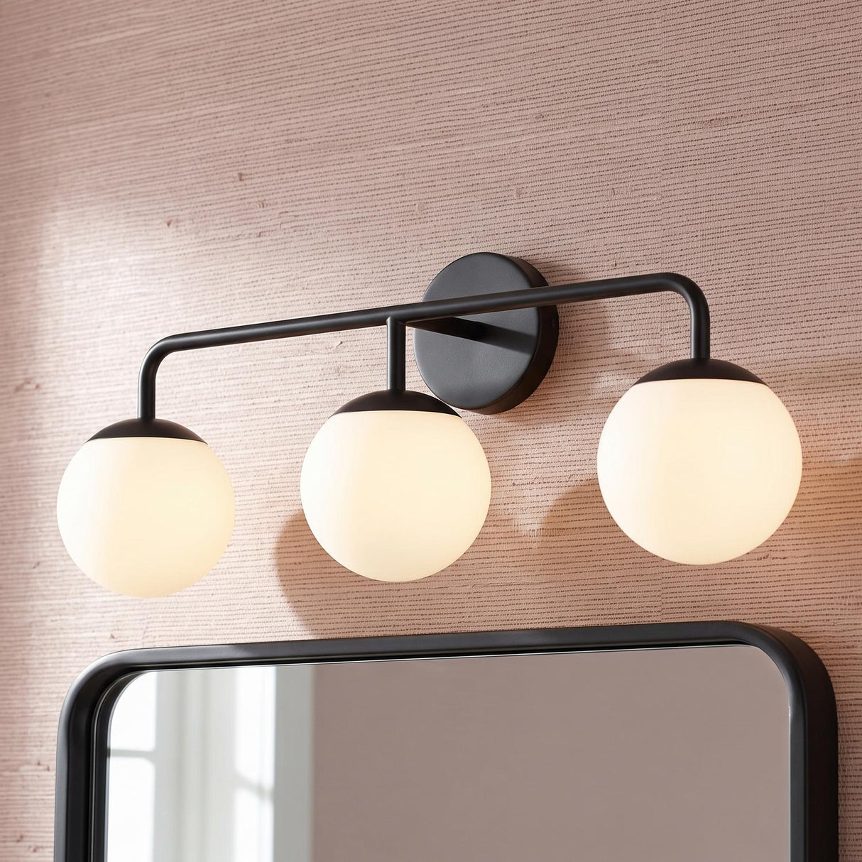 A modern bathroom with the Castell 3 Globe Vanity, Matte Black featuring three frosted glass globes above a black framed mirror and textured pink grasscloth wallpaper.