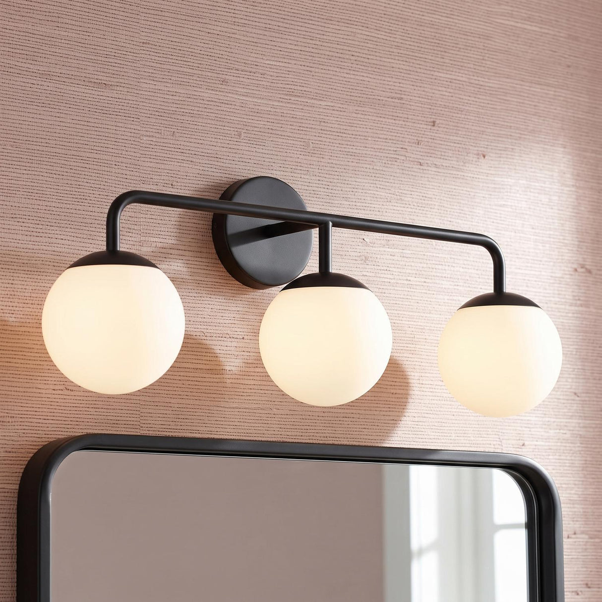 A modern bathroom with the Castell 3 Globe Vanity, Matte Black featuring three frosted glass globes above a black framed mirror and textured pink grasscloth wallpaper.