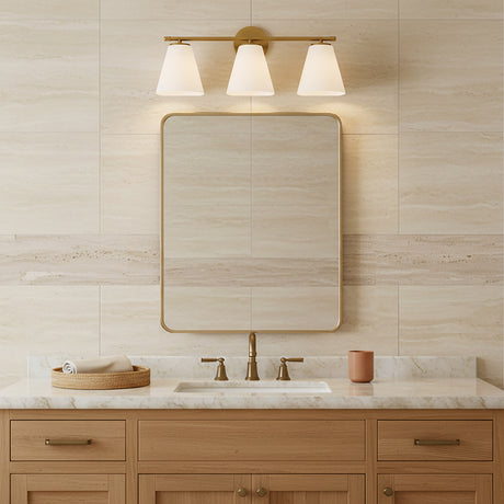 Carlisle 3 Light Vanity, Brushed Brass with Opal Glass - A vanity light fixture with three opal glass shades in a brushed brass finish is mounted above a framed mirror in a bathroom with travertine-style tile.