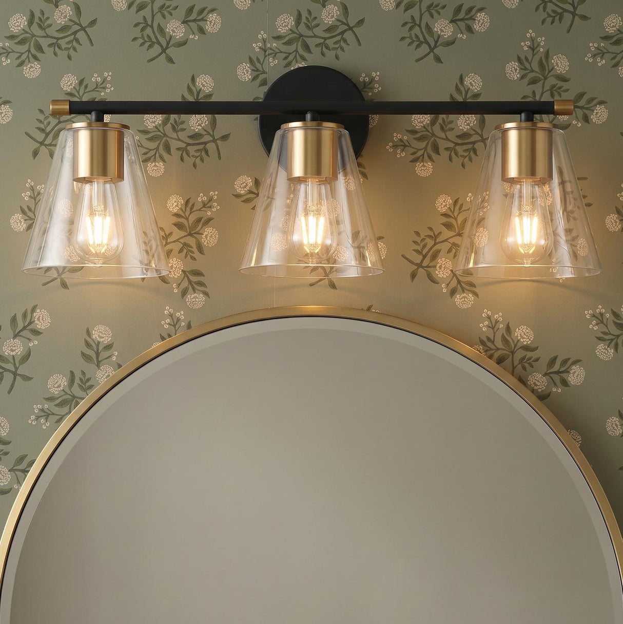 A transitional bathroom featuring the Carlisle 3 Light Vanity, Matte Black and Brushed Brass, installed above a gold round mirror against green floral wallpaper.
