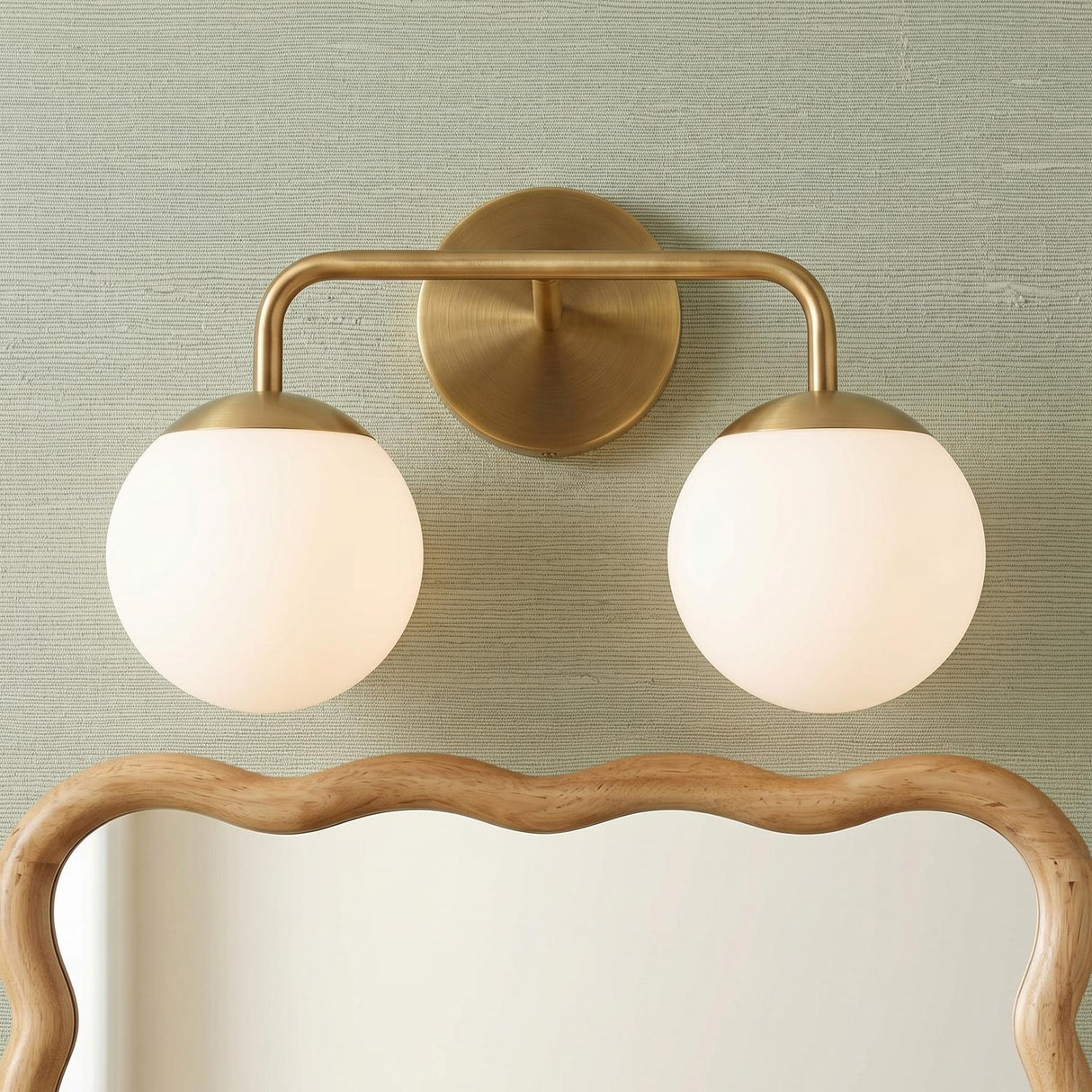 A modern bathroom with the Castell 2 Globe Vanity Light, Aged Brass featuring opal glass globes mounted above a wavy wood-framed mirror on sage green textured wallpaper.