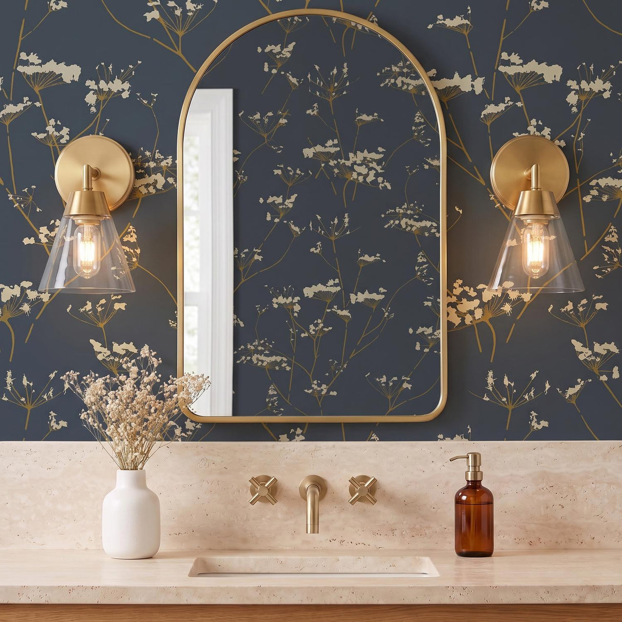 A transitional bathroom with two satin brass and clear glass wall sconces flanking an arched gold mirror against navy botanical wallpaper and travertine counters.