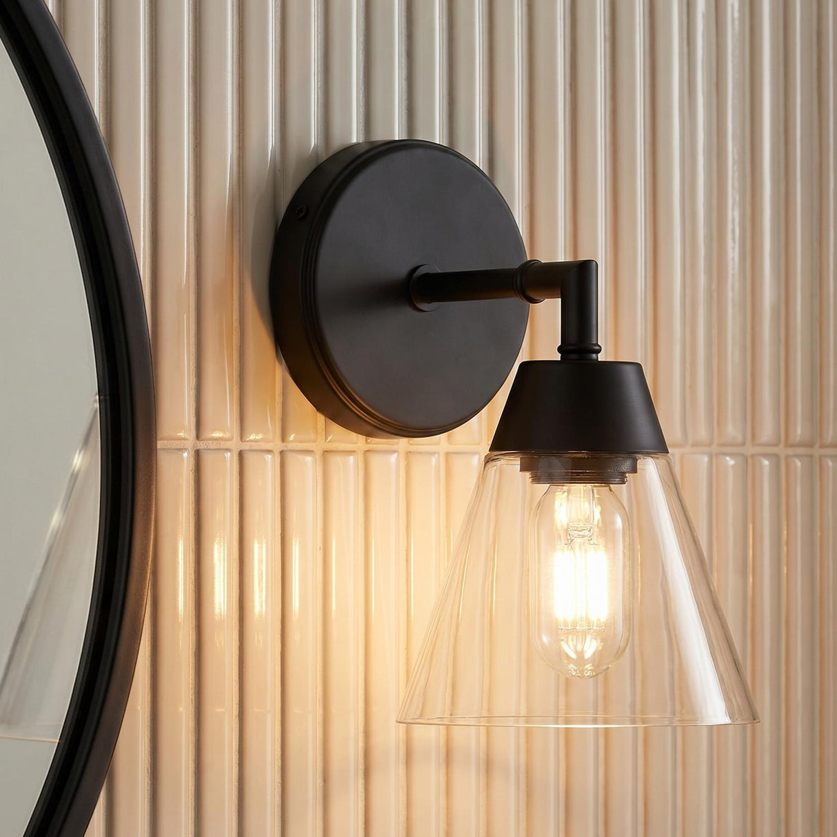 A modern bathroom with an Owen 1 Light Vanity Wall Sconce, Matte Black featuring a tapered glass shade mounted on fluted cream tiles beside a round mirror.