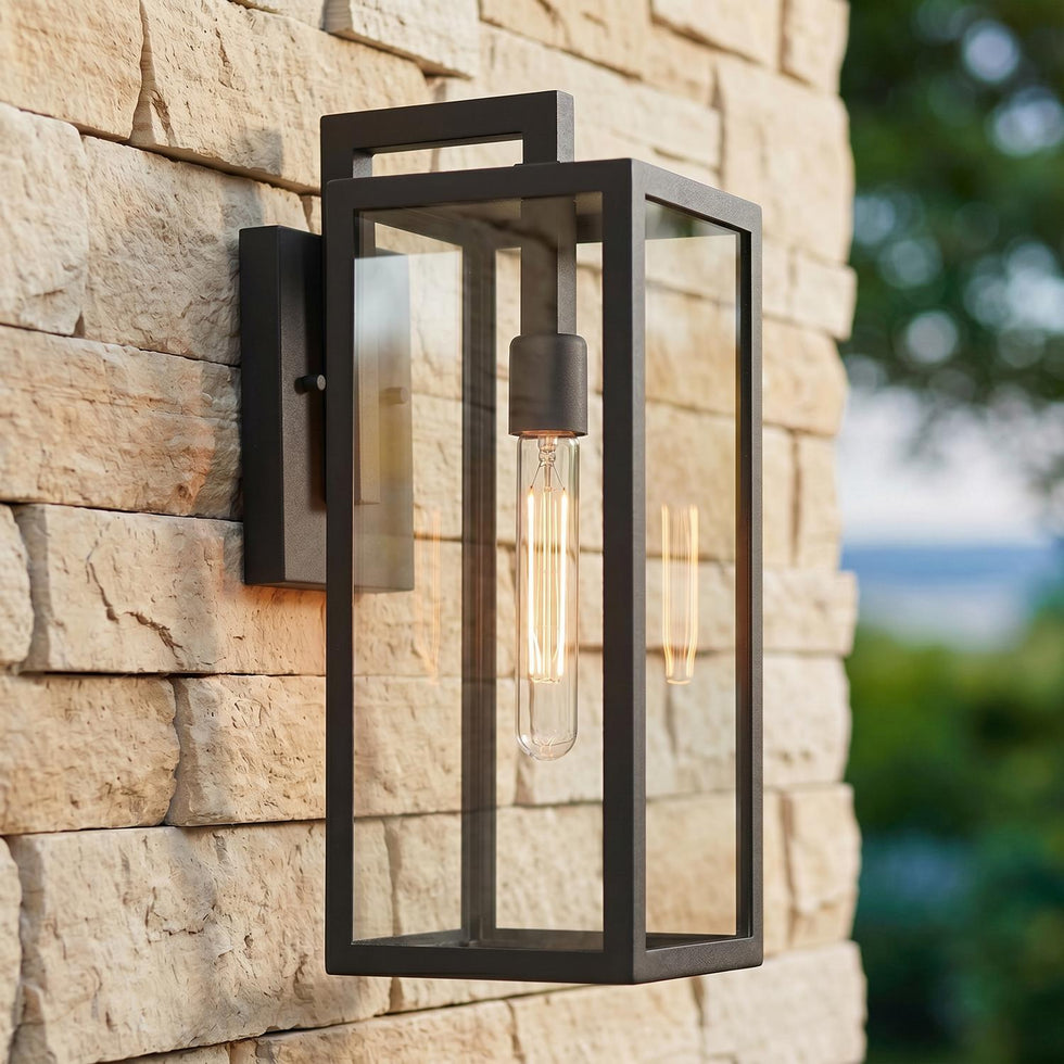 A rectangular modern home exterior with a textured black Sam Outdoor Wall Light, Medium 18" glowing on a tan stacked stone wall beside blurred green landscaping at twilight.