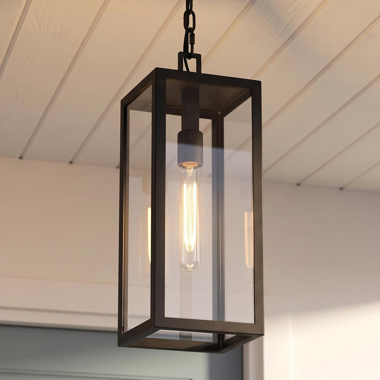 A modern farmhouse porch with the Sam Outdoor Hanging Light in matte black suspended from a white shiplap ceiling with a glowing Edison bulb at dusk.