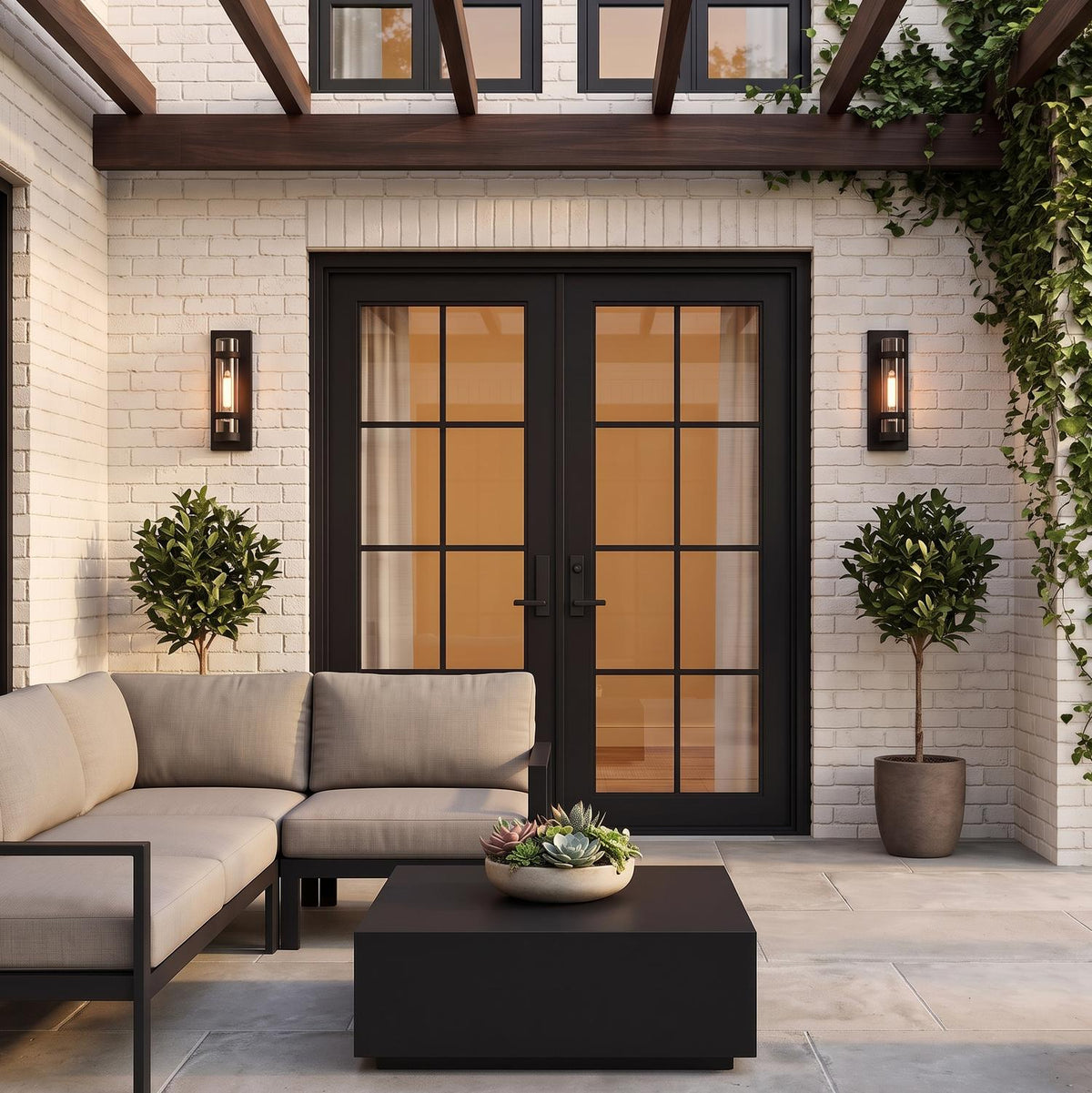 A modern farmhouse patio with two Glenn Outdoor Wall Light, Matte Black lanterns flanking black-framed glass doors on white brick at dusk, featuring potted boxwood trees.