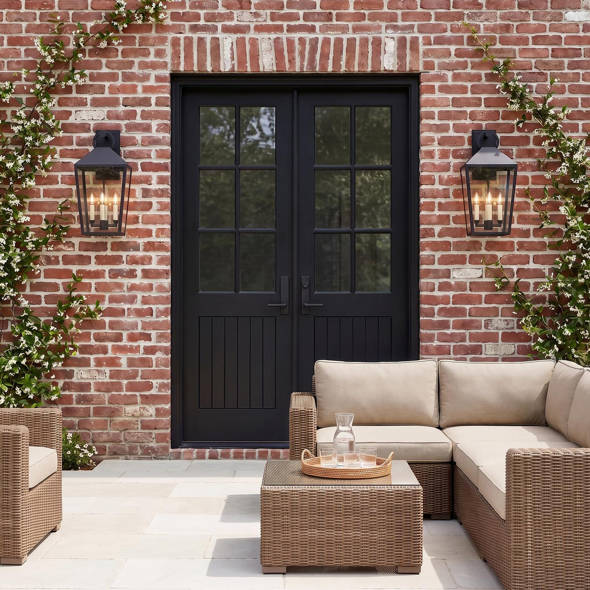 A traditional red brick patio features two matte black Cole Outdoor Wall Light lanterns flanking black French doors at twilight, surrounded by climbing jasmine and wicker seating.