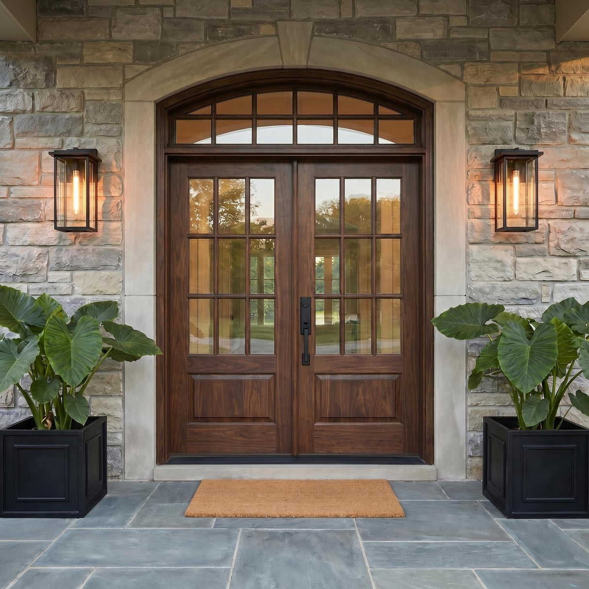 A transitional home entry with two matte black wall lanterns flanking dark wood double doors on a stone facade at twilight with large potted elephant ear plants.
