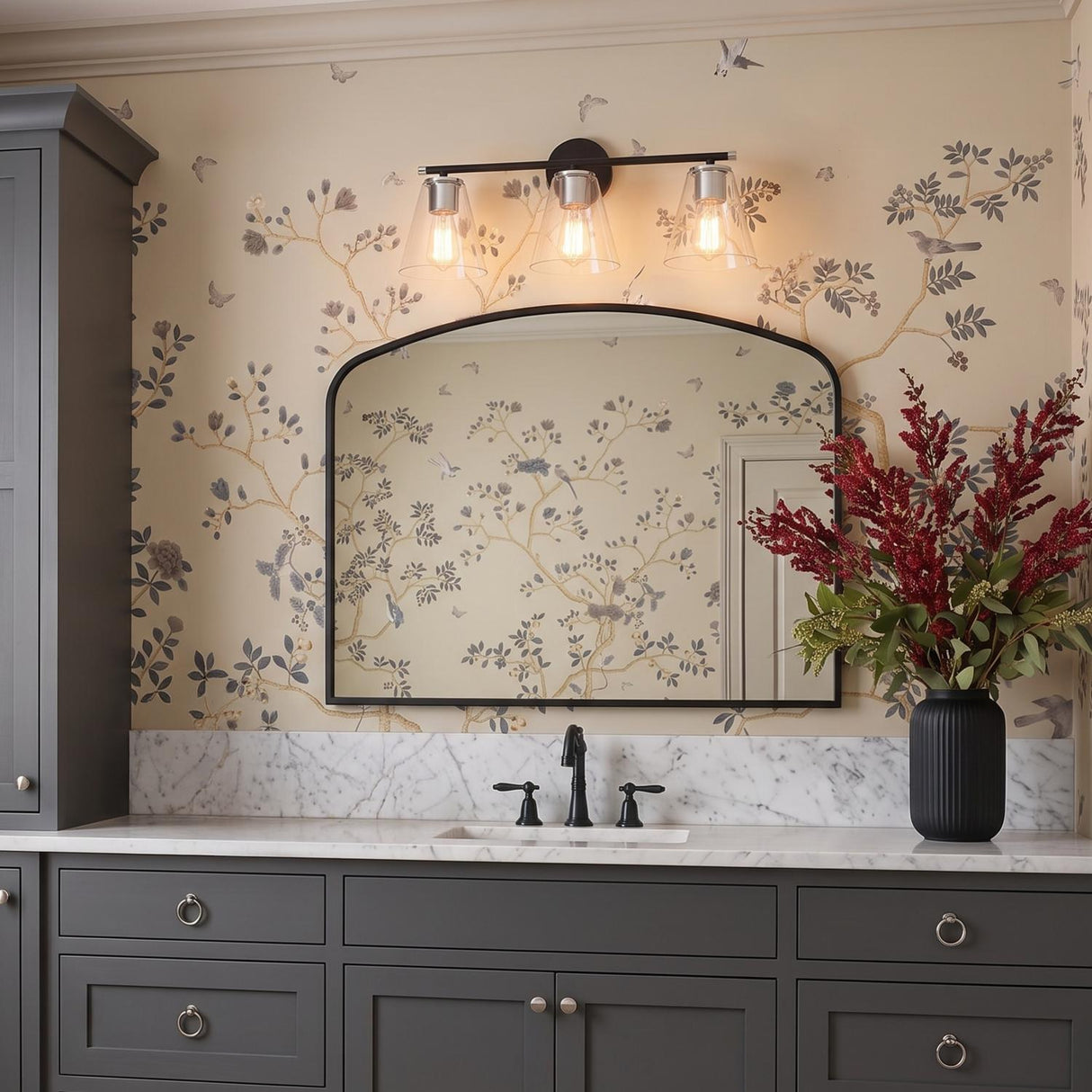 A traditional bathroom with the Carlisle 3 Light Vanity, Matte Black and Chrome, installed above an arched mirror, white marble countertop, and grey shaker cabinets with floral wallpaper.