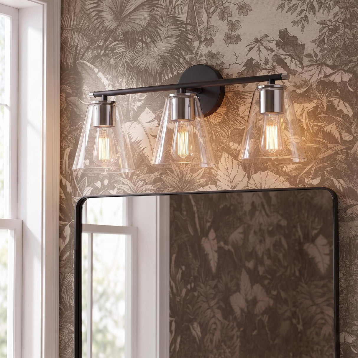 A transitional bathroom featuring a matte black and chrome vanity light with tapered glass shades mounted above a black mirror and sepia-toned botanical wallpaper.