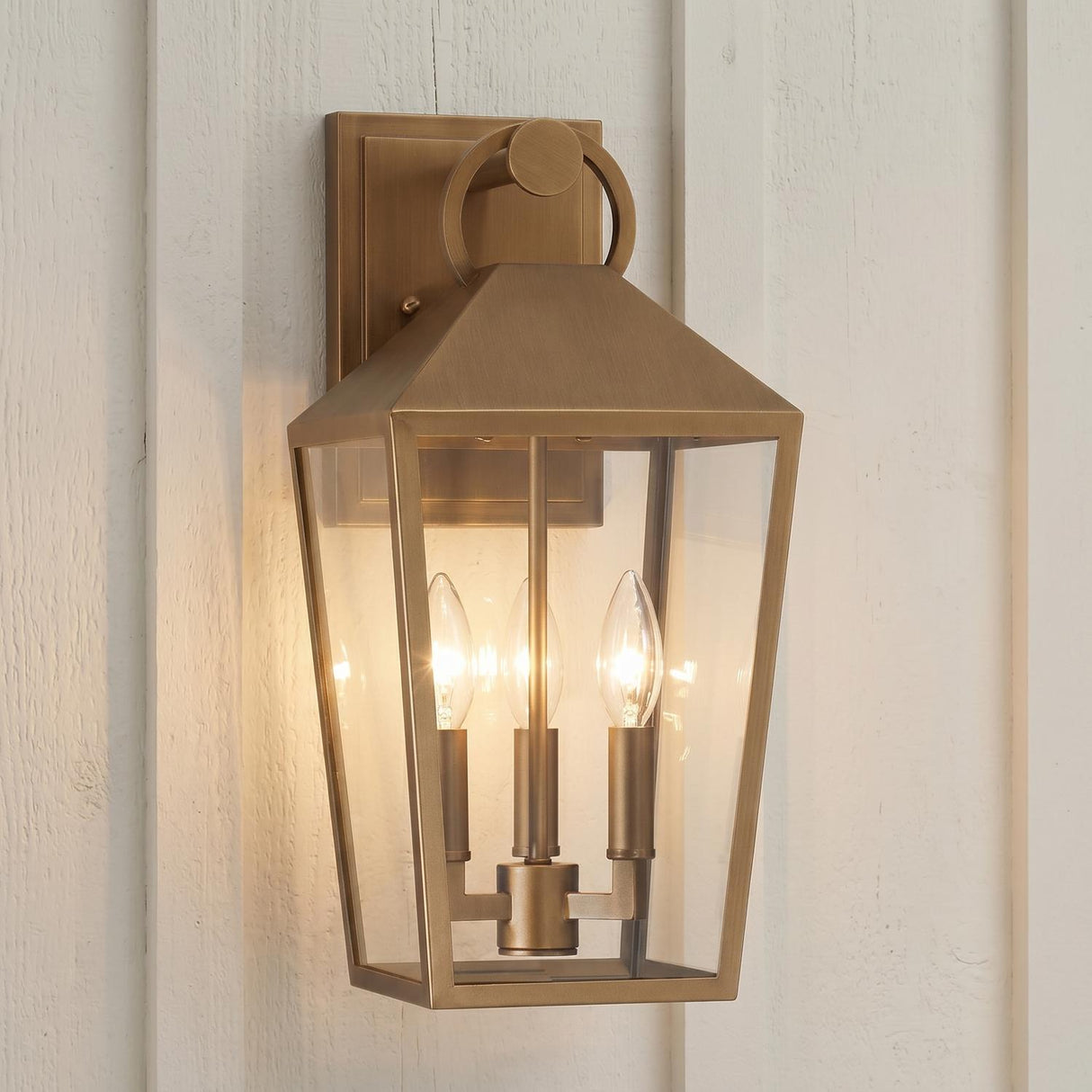 An outdoor entryway featuring the Cole Outdoor Wall Light, Medium 17 inch Burnished Brass lantern with clear glass panels mounted against white board and batten siding.