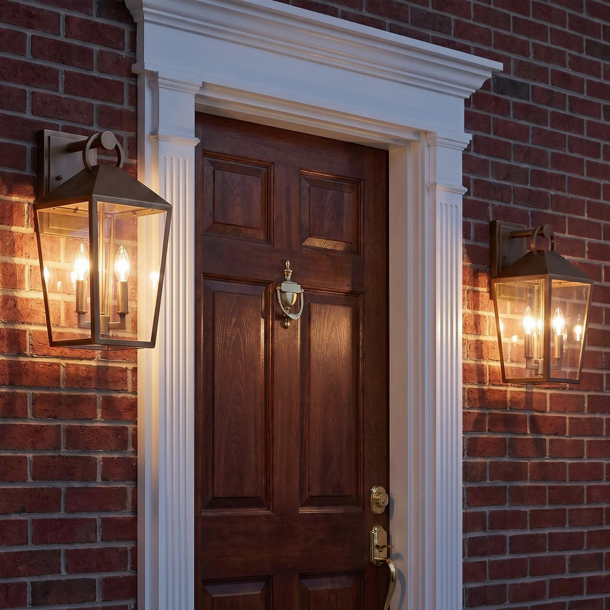 A traditional brick entryway featuring two Cole Outdoor Wall Lights in burnished brass flanking a dark wood door with white decorative trim and a brass knocker.