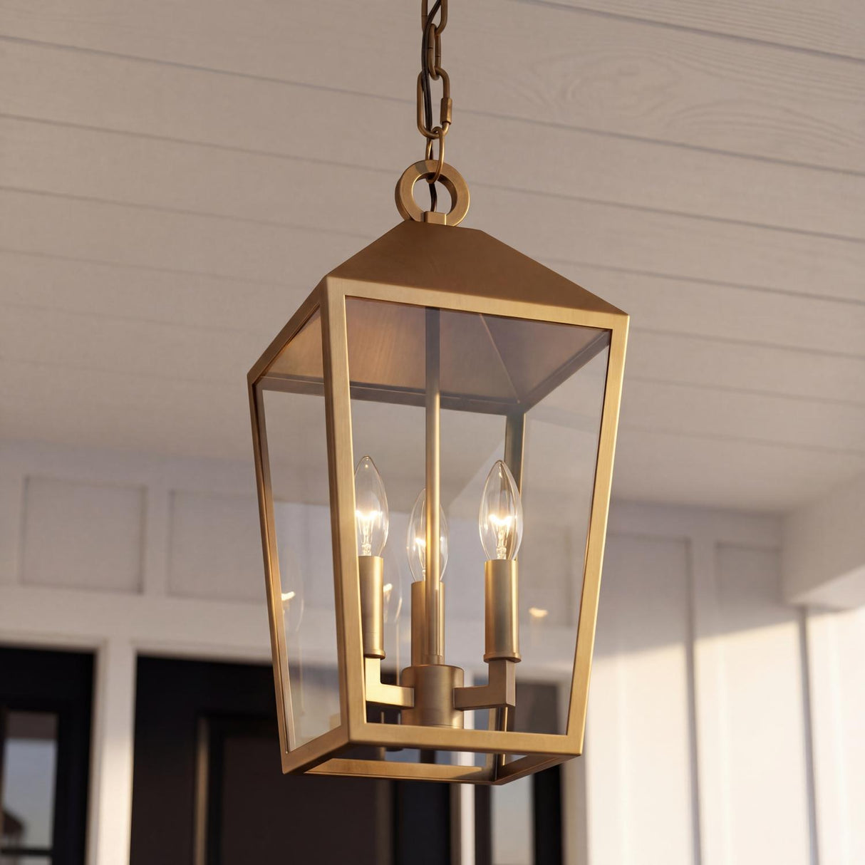 An outdoor porch with a Cole Outdoor Hanging Light, Burnished Brass with clear glass panels suspended from a white shiplap ceiling in front of a black door.