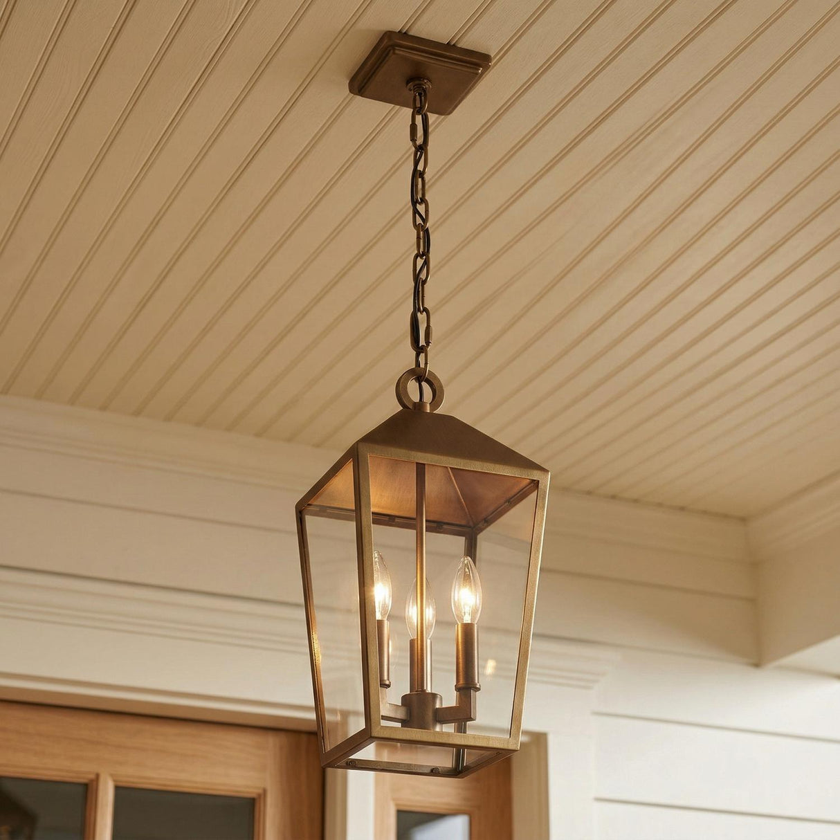 An outdoor porch featuring the Cole Outdoor Hanging Light, Burnished Brass lantern suspended from a cream beadboard ceiling above a wood door with white shiplap siding.