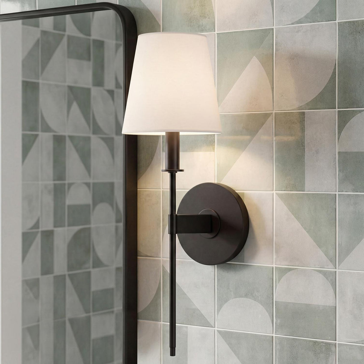 A contemporary bathroom features a Taylor Wall Sconce, Matte Black with a white linen shade installed on a geometric sage green tiled wall beside a mirror.