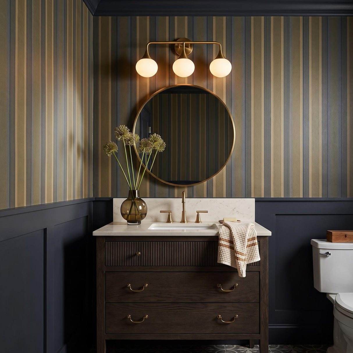 A traditional bathroom featuring the Talia 3 Light Vanity, Aged Brass with frosted glass globes over a round mirror, marble countertop, and striped wallpaper.