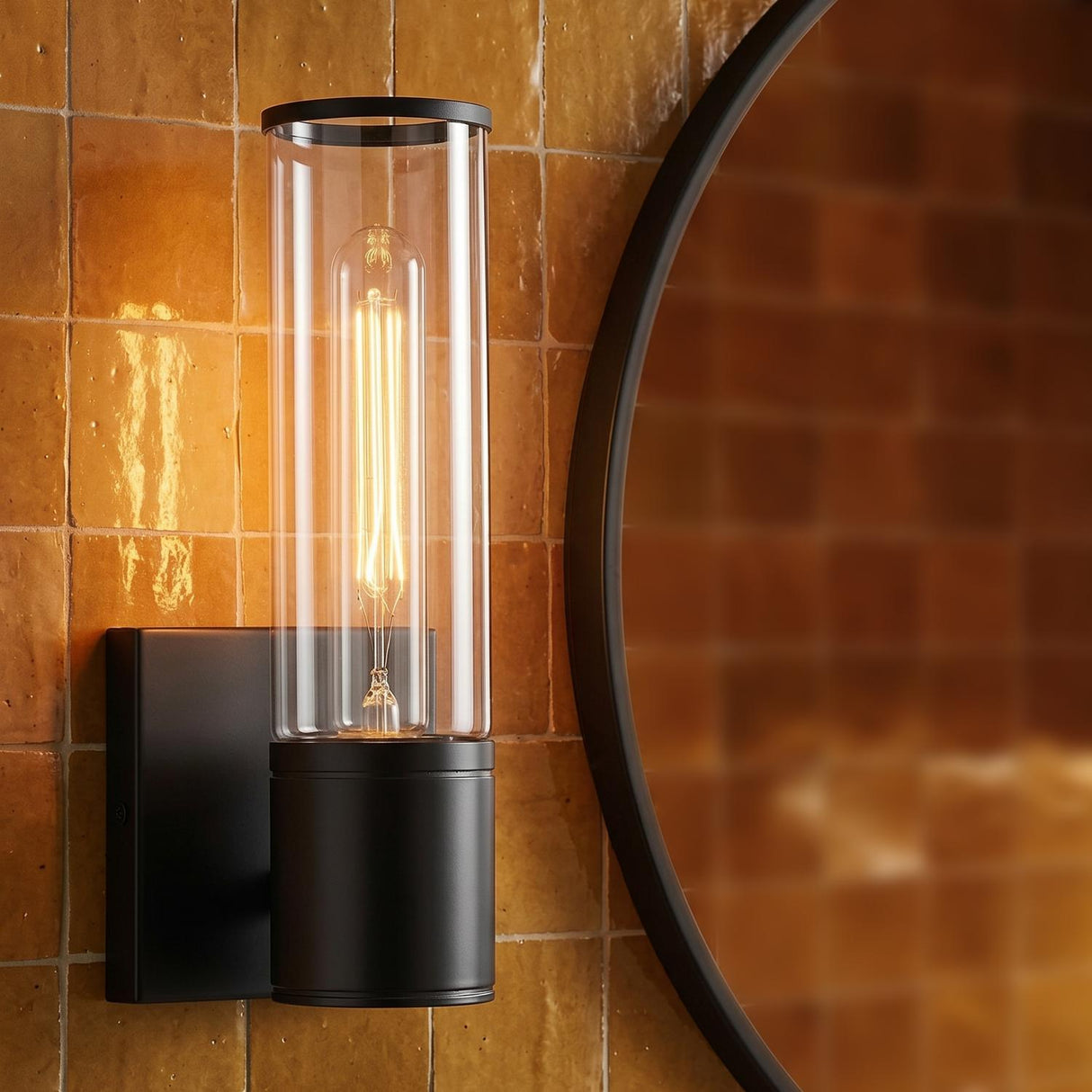 A contemporary bathroom with a matte black Lea Wall Sconce featuring a clear glass cylinder mounted on warm terracotta tile beside a circular black mirror.