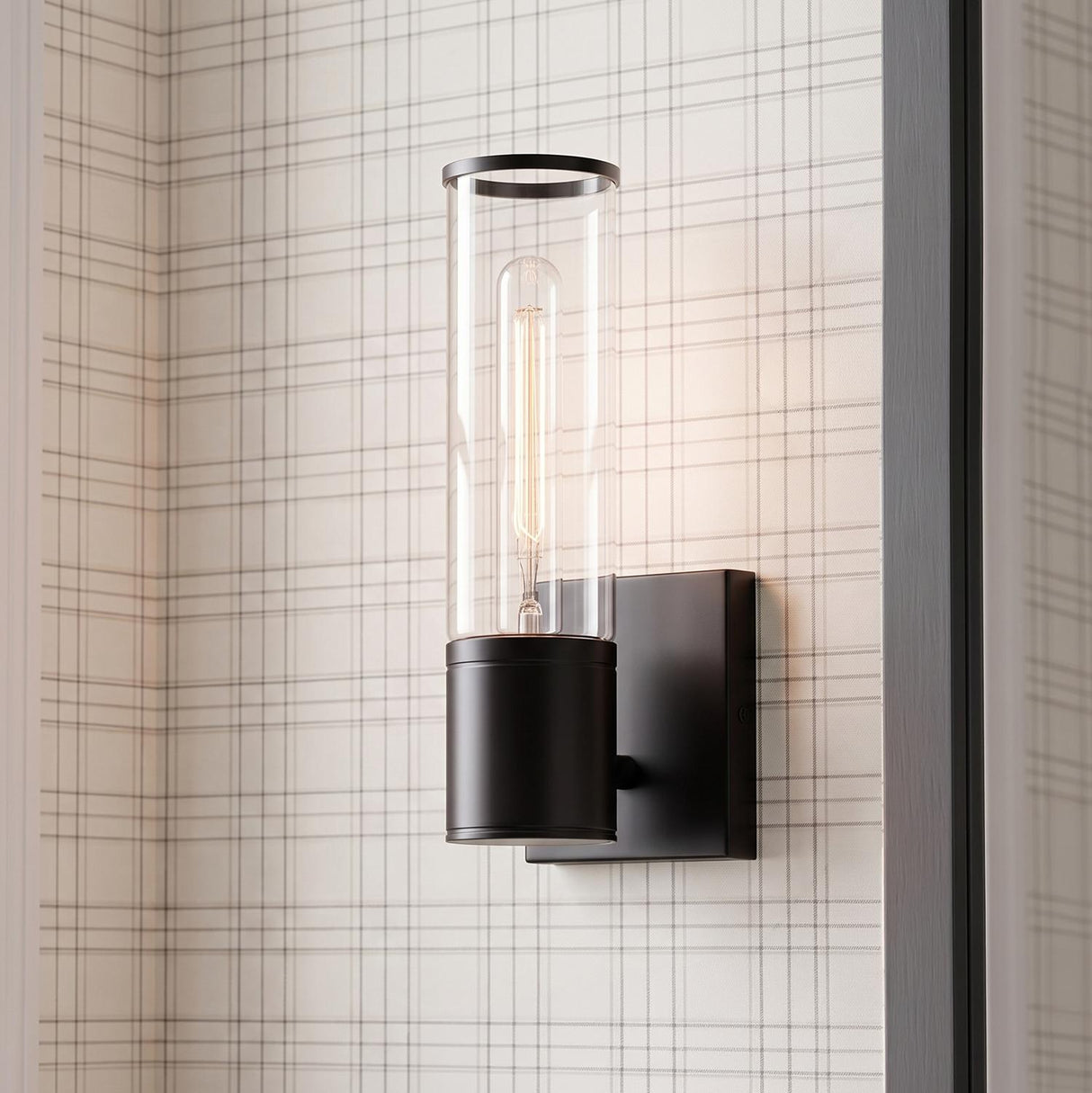 A modern farmhouse foyer featuring the Lea Wall Sconce, Matte Black with a clear glass cylinder and Edison bulb mounted on neutral plaid wallpaper beside a doorway.