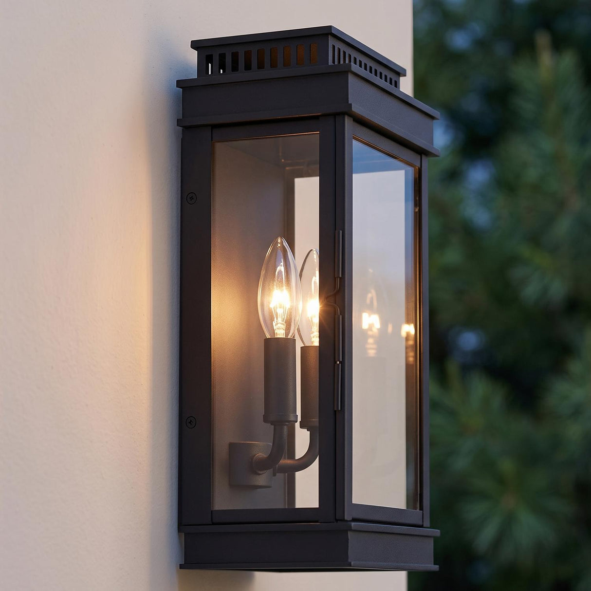 A modern home exterior featuring a matte black wall lantern glowing on white stucco at dusk, with the Brighton Outdoor Wall Light, Black beside evergreen trees.