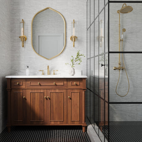 Camie 20 inch tall art-deco wall sconce with reeded glass and aged brass finish in am updated vintage bathroom, flanking a brass vanity mirror with marble tiled walls.