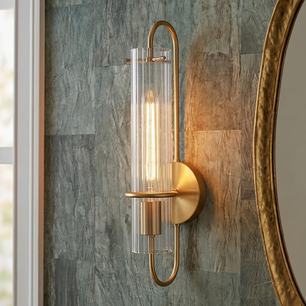 A contemporary bathroom featuring the Camie Wall Sconce 20", Aged Brass with ribbed glass mounted on a textured green wall beside a round gold mirror.
