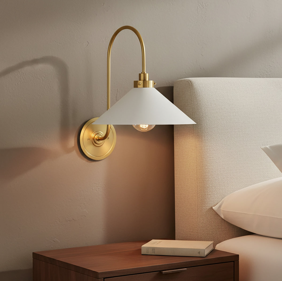Alma wall sconce in aged brass finish with white shade above a nightstand next to a bed.