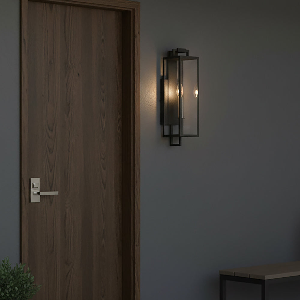 Everett Outdoor Wall Light, Small, Forged Iron Black - A modern outdoor wall light with a black metal frame and clear glass panels is mounted on a gray wall next to a wooden door.