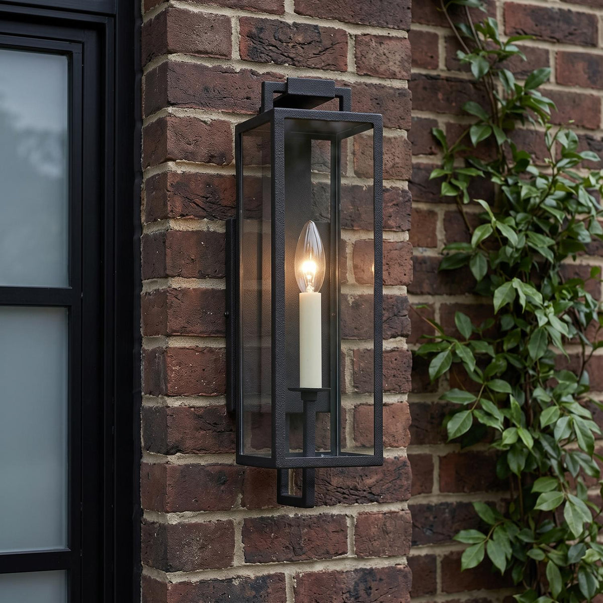 A modern brick home exterior with the Everett Outdoor Wall Light in forged iron black glowing on a red brick wall beside green climbing vines at dusk.