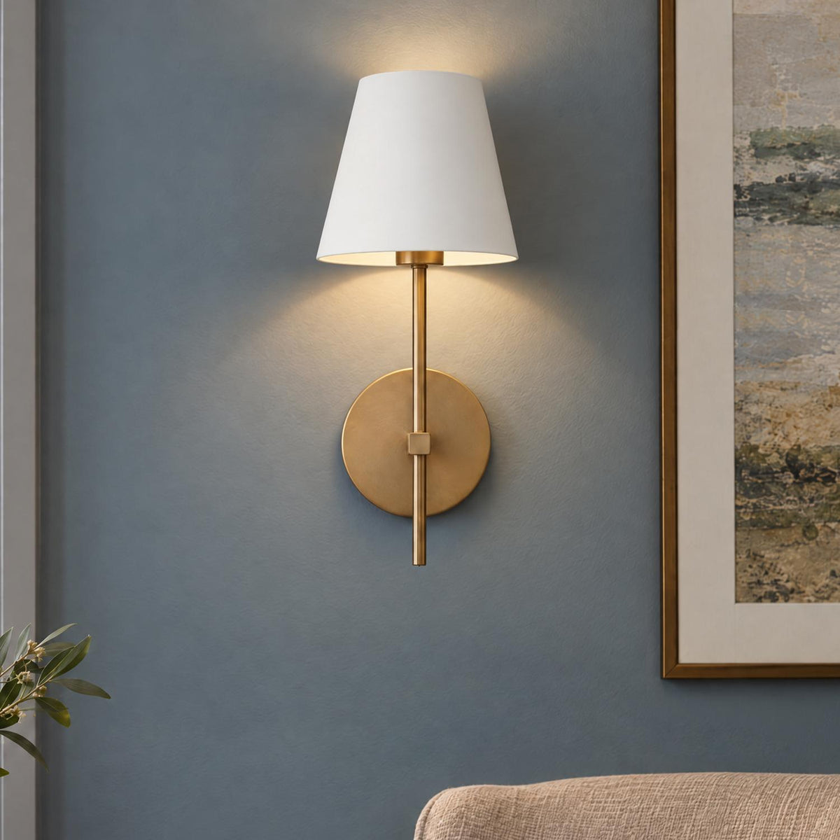 A transitional living room featuring a Banks Wall Sconce, White and Brass with a tapered fabric shade mounted on a blue wall beside framed artwork.