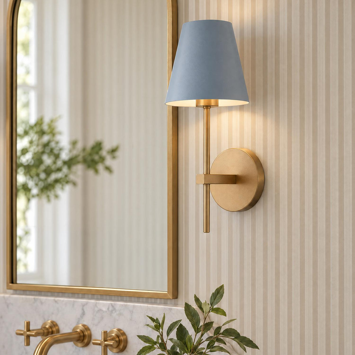 A transitional bathroom vanity features the Banks Wall Sconce, French Blue and Brass, mounted beside a brass arched mirror above a white marble countertop with striped wallpaper.