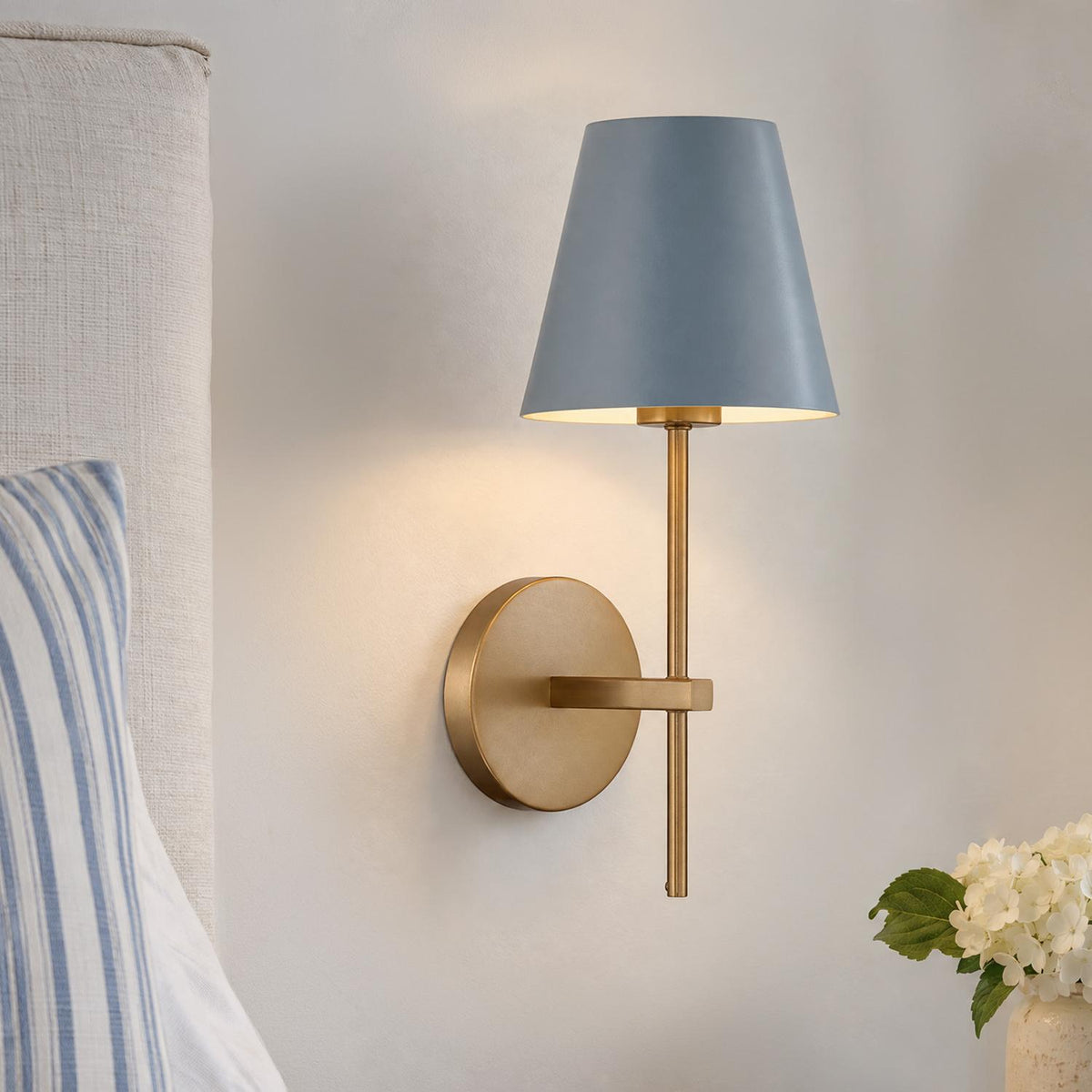 A transitional bedroom featuring the Banks Wall Sconce, French Blue and Brass mounted beside a neutral upholstered headboard with a striped blue pillow.