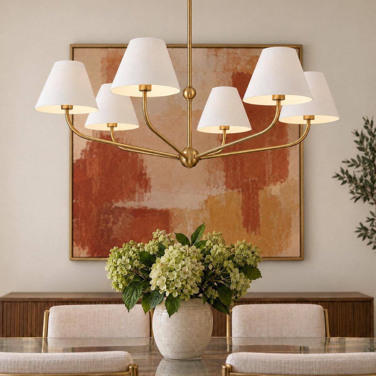 A transitional dining room features a brushed brass chandelier with white fabric shades suspended above a glass dining table with a green hydrangea centerpiece.