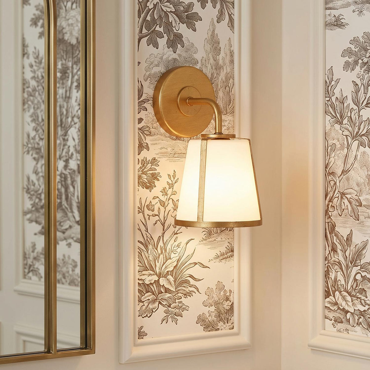 A traditional bathroom features the Hoyt Vanity Wall Sconce, antique gold with opal glass, mounted on a brown botanical toile wallpaper panel beside an arched mirror.
