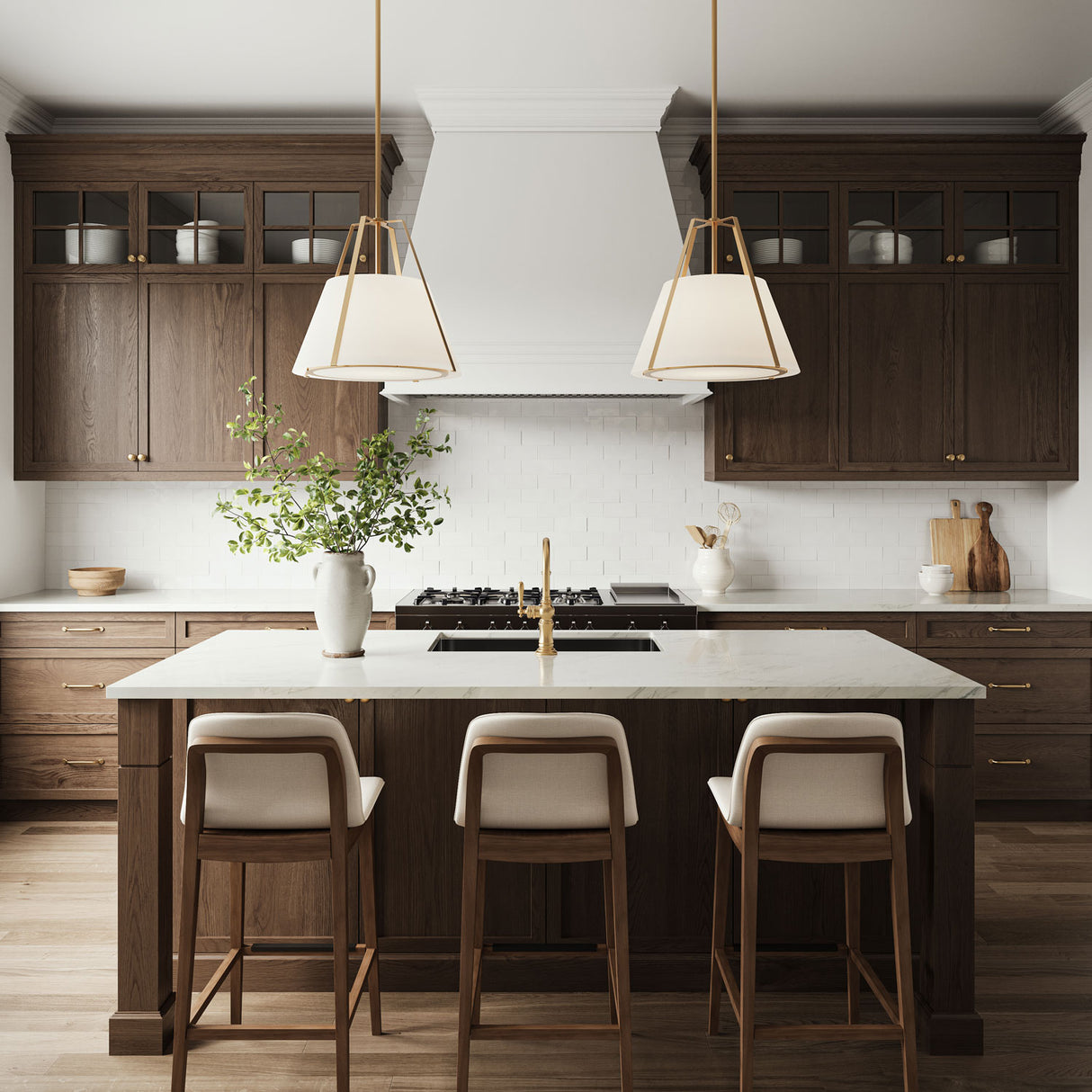Two 20" wide Hoyt Pendant lights in Antique Gold with white shades are shown in a traditional kitchen, hung over a kitchen island, with dark walnut cabinetry and bright white stone countertops, with seating for three.