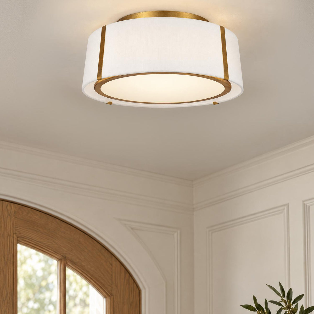 A transitional foyer features an antique gold flush mount light with a white fabric drum shade installed on the ceiling near an arched wood door.