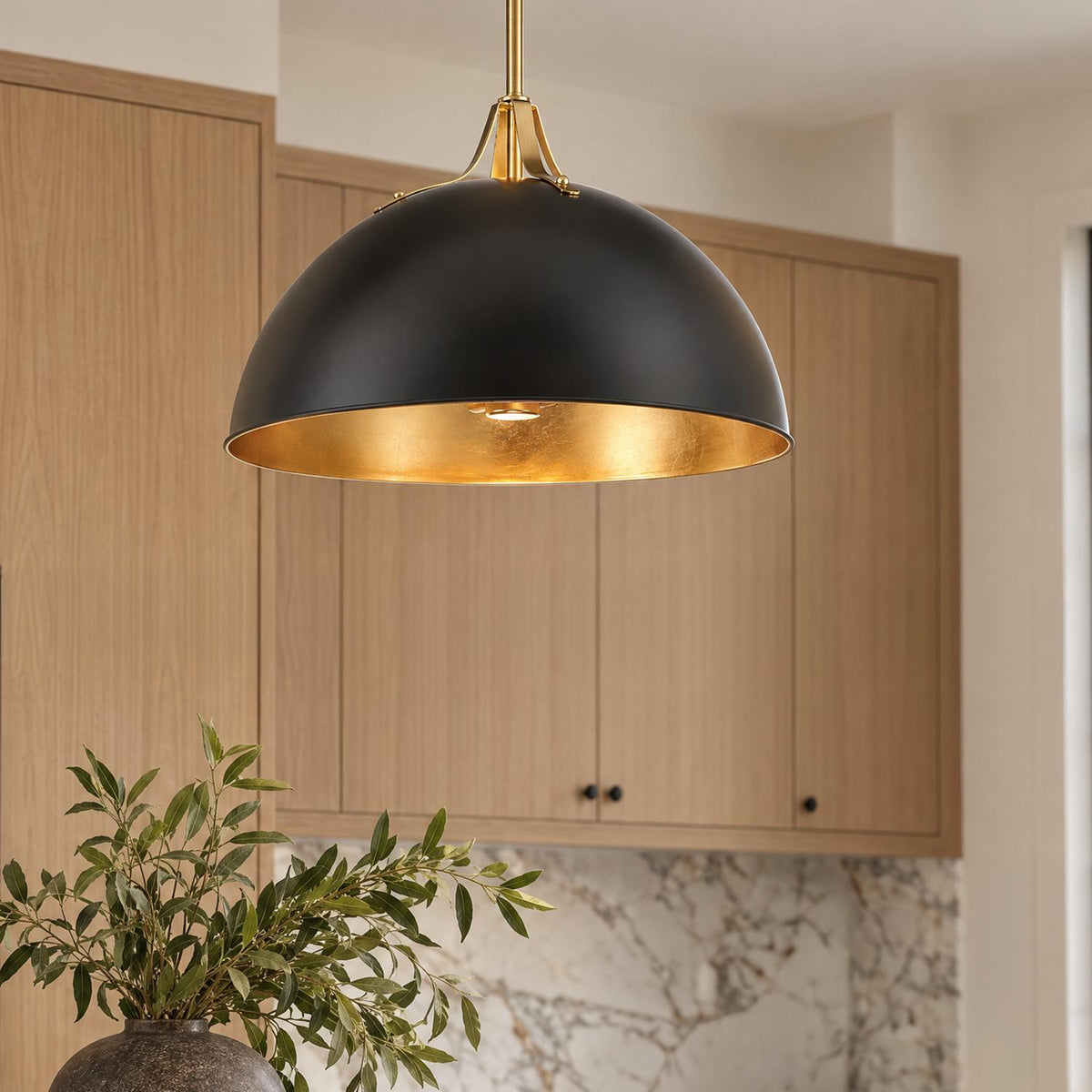 A modern kitchen features the Grant Extra Large Pendant 23", Black and Antique Gold hanging in front of light wood cabinets and marble backsplash.