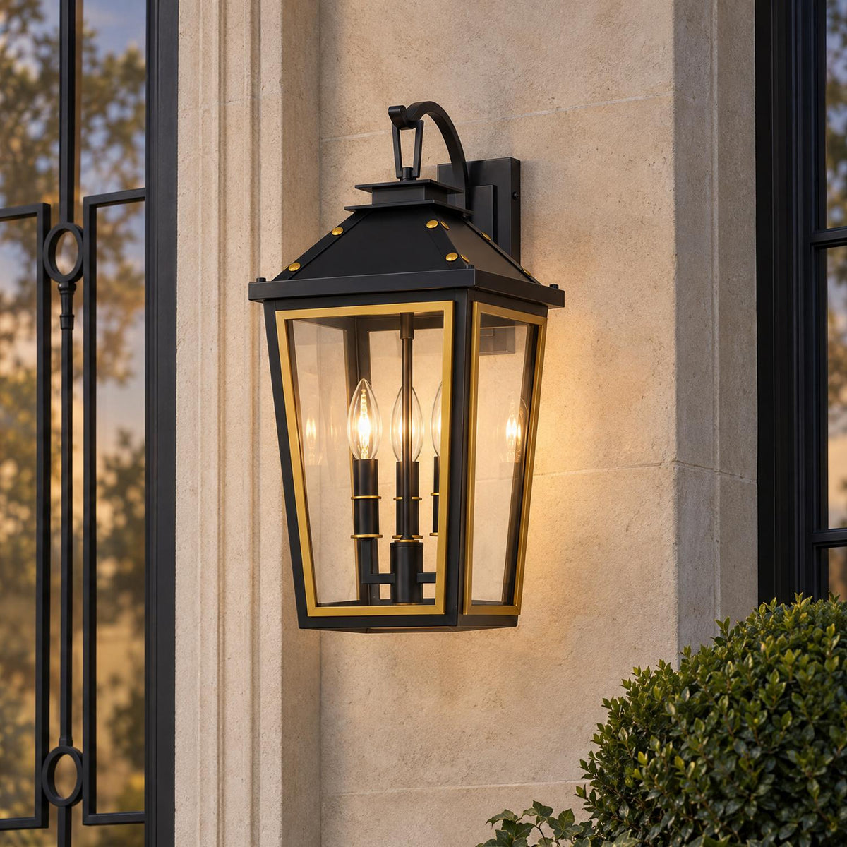 A transitional home exterior with the glowing Ellington Outdoor Wall Light, Medium 19" Black and Textured Gold mounted on a limestone wall beside boxwood greenery at dusk.