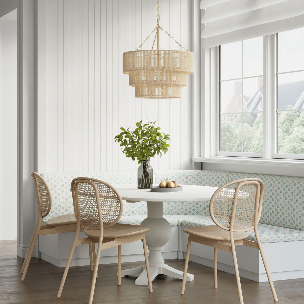 Dining area with round table and wicker chairs in a bright room with large windows, hanging above is the Sorelle 10 Light chandelier with soft brass hardware.