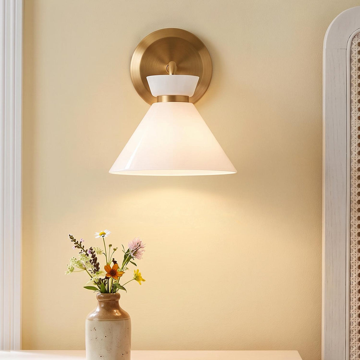 A mid-century bedroom features the Cori Wall Sconce, Brushed Brass and Milk Glass mounted on a beige wall beside a woven cane headboard and fresh wildflowers.