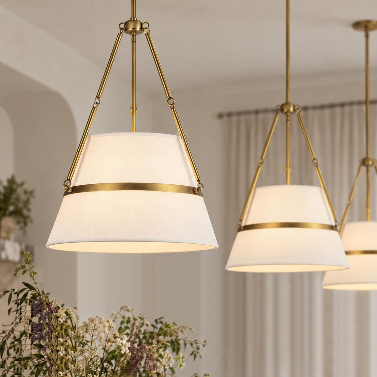 A transitional dining room featuring three antique brass pendants with white linen shades hanging in a row near beige drapes and a fresh floral arrangement.