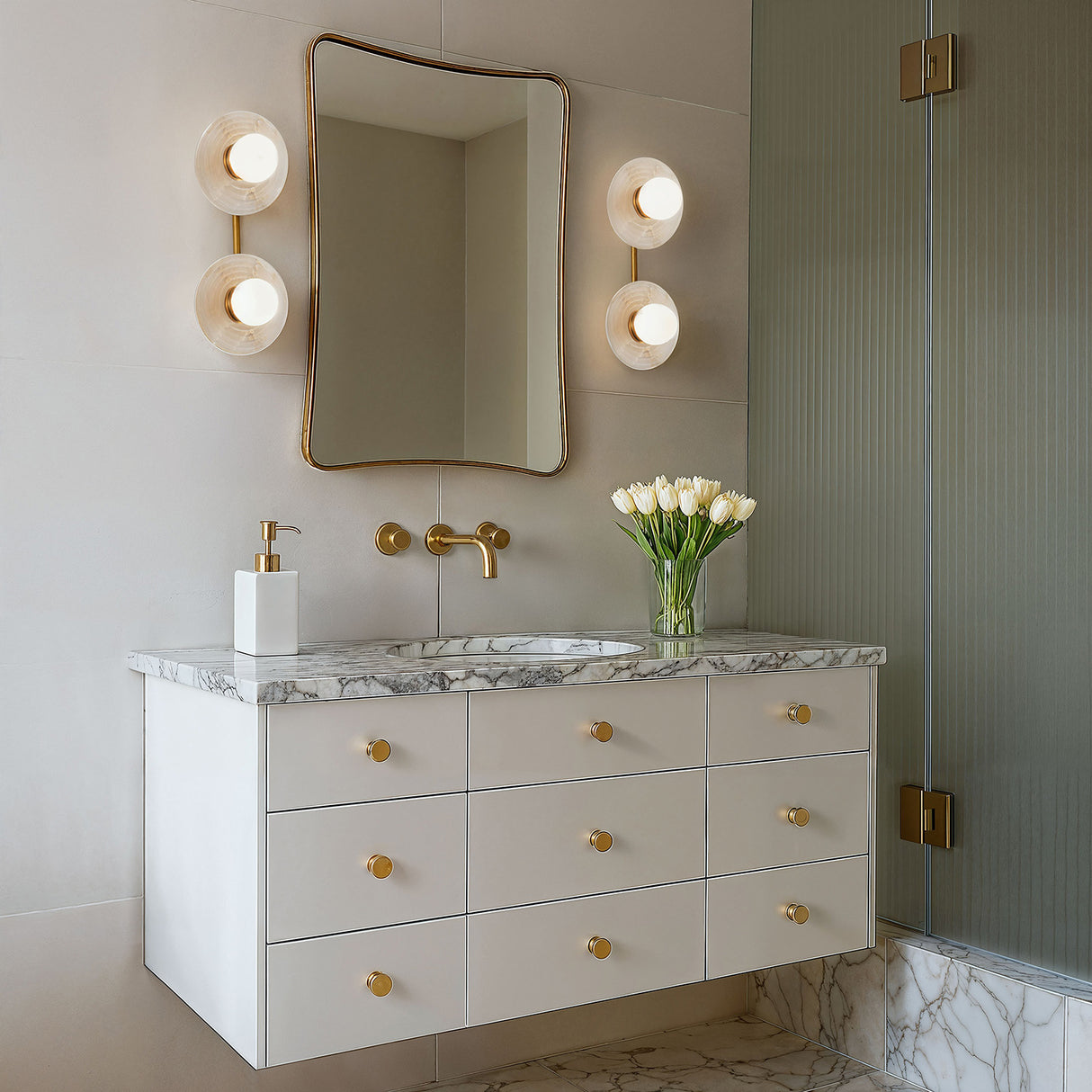 The Bren 2 Light Wall Sconce in Vinatge Brass with natural Alabaster tone is shown in a contemporary bathroom, flanking a curved brass mirror above a floating vanity.