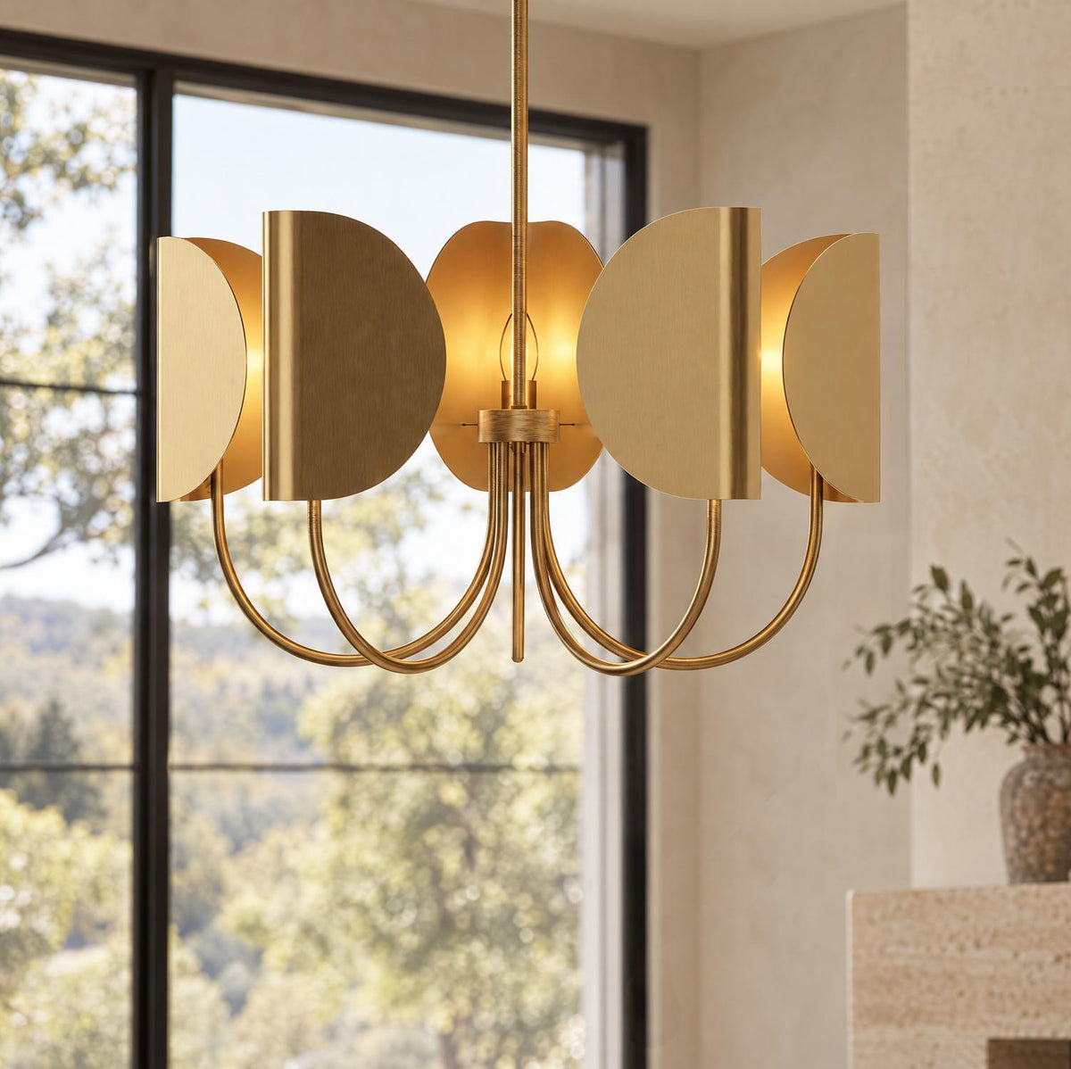 A contemporary living room showcases the Nico 5 Light Chandelier 32", Antique Brass positioned beside a large black-framed window with scenic outdoor views.