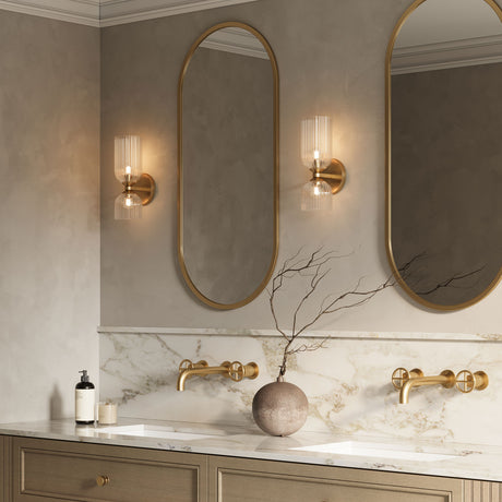 Elle Wall Sconce shown in brass with clear glass in a bathroom, flanking tall oval shaped vanity mirrors.